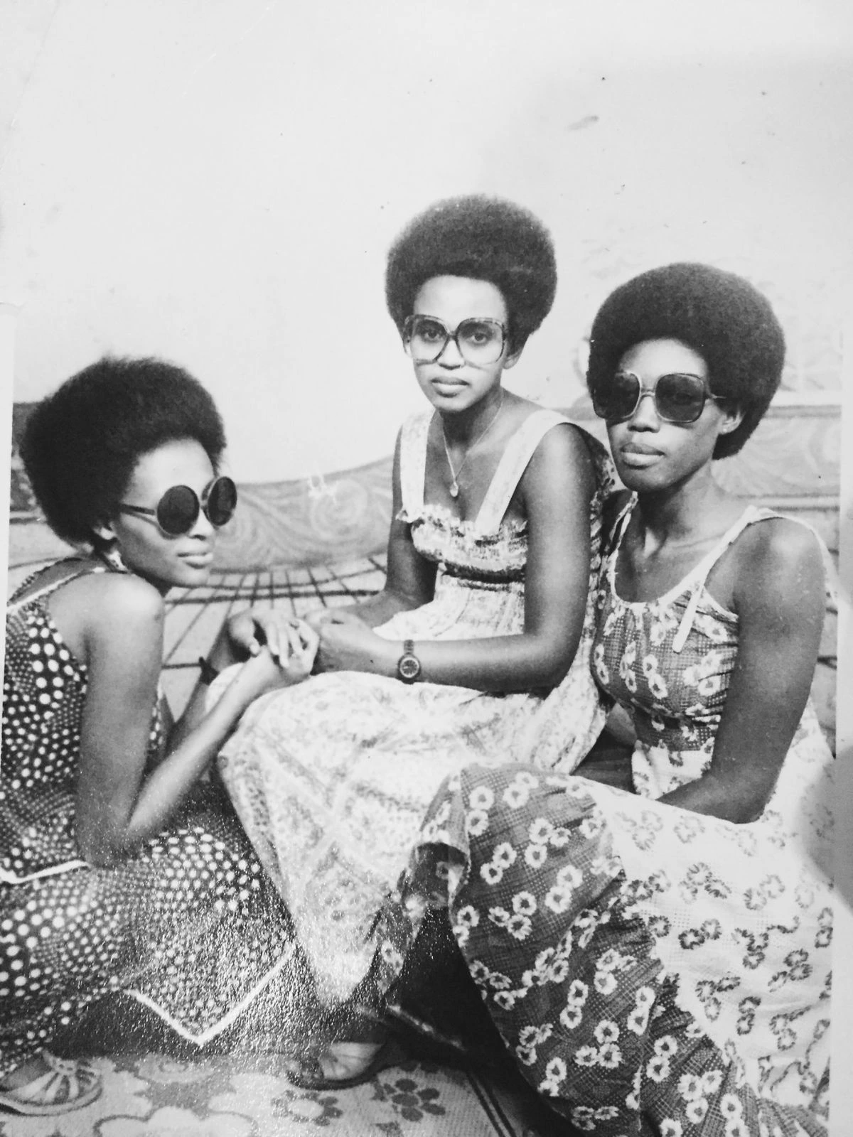 Three young Nigerian women in the 1970s sporting afro hairstyles, oversized tinted sunglasses, and patterned sundresses.