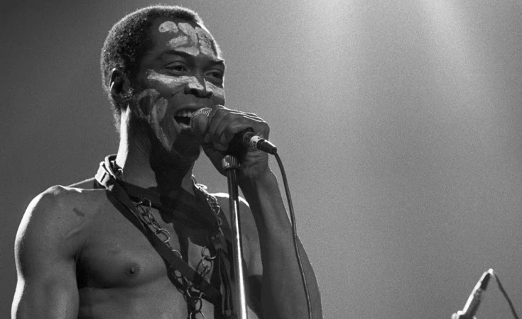 Afrobeat pioneer Fela Anikulapo Kuti singing intensely with white clay face markings during a live concert.